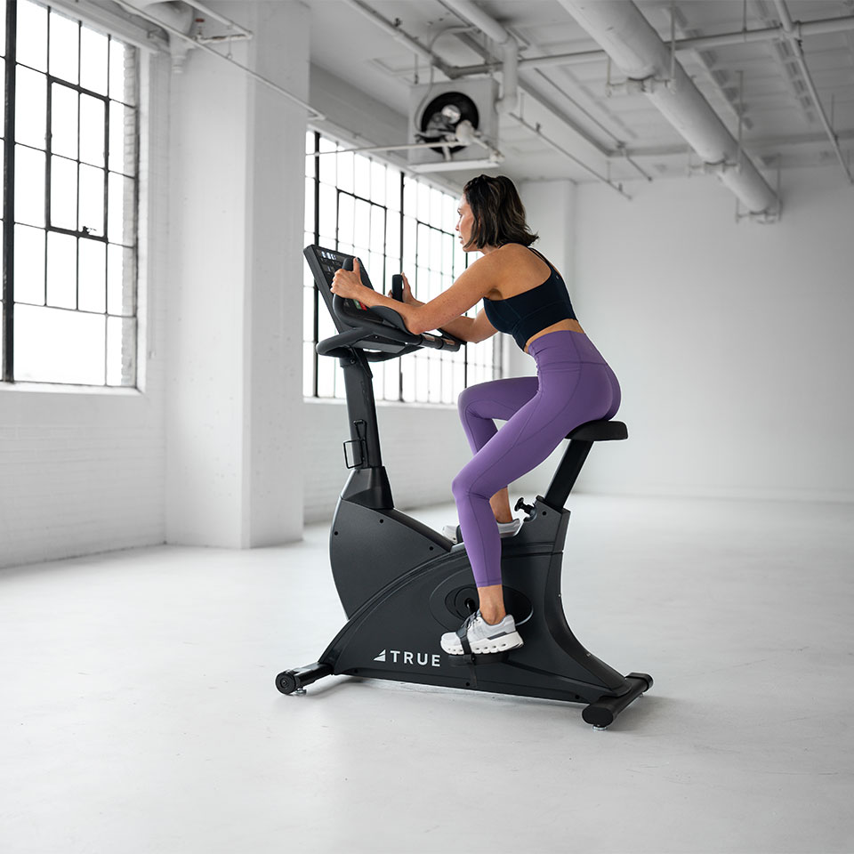 A woman in a black sports bra and purple leggings rides a stationary exercise bike in a bright, spacious room with large windows and white walls.