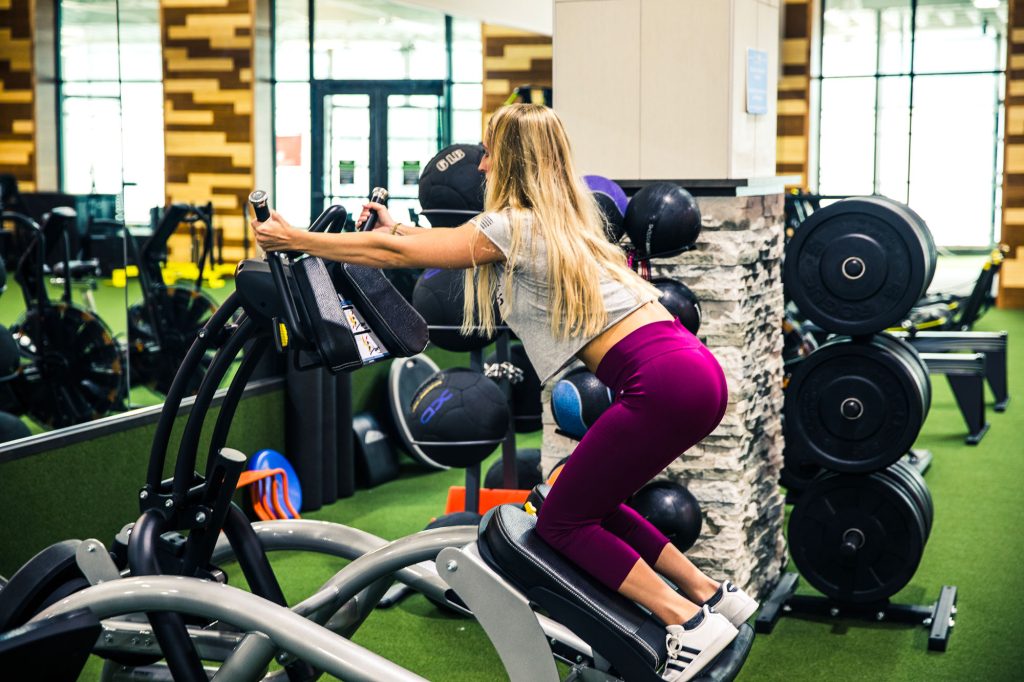 A woman with long blonde hair and a gray top uses a gym machine for leg and glute exercises in a modern fitness center with weights and equipment in the background.