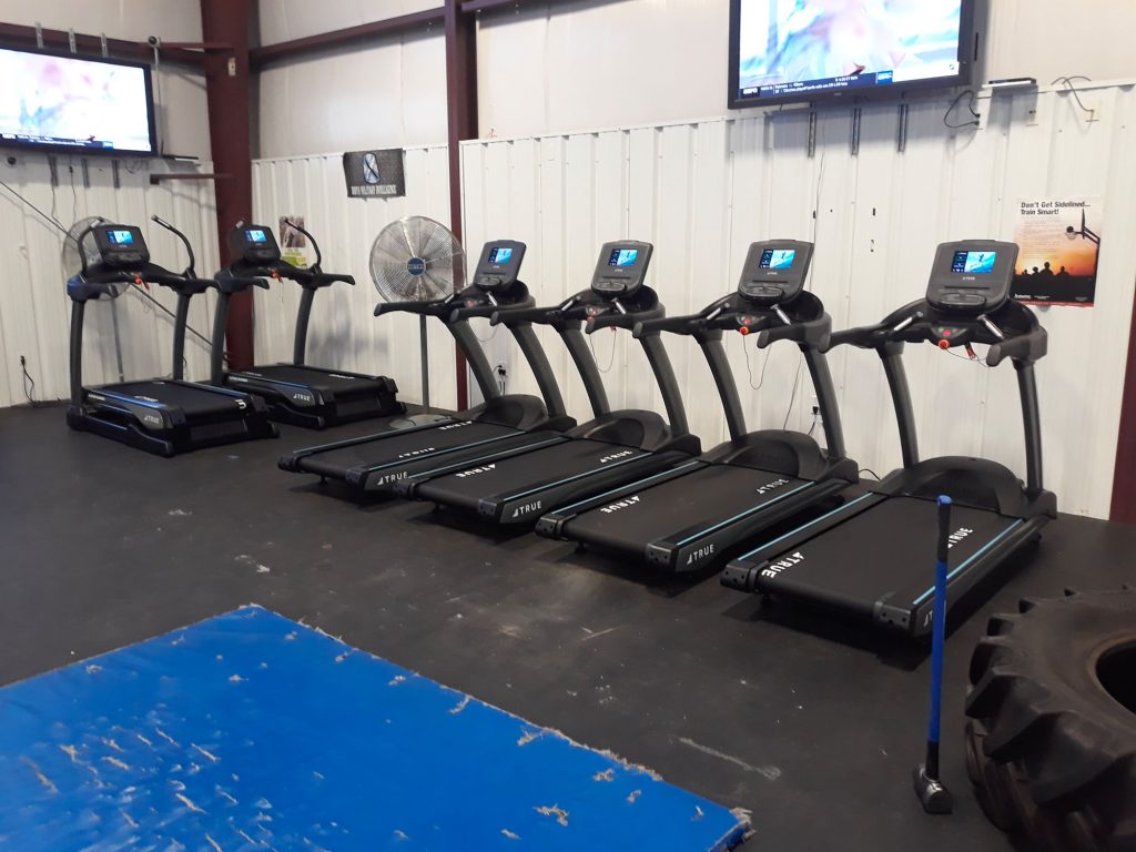 Six treadmills are lined up in a gym with a blue mat in the foreground, a large fan on the back wall, TV screens mounted above, and a large tire on the right side. The floor and walls are industrial style.
