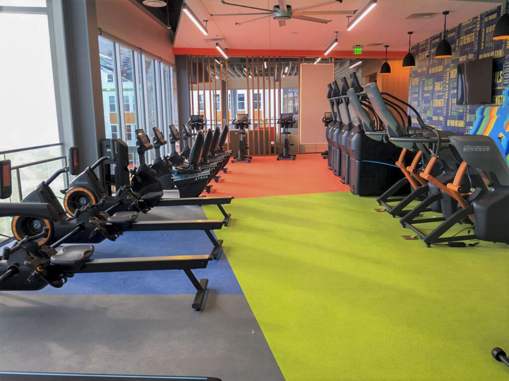 A modern gym with rowing machines lined up on the left and treadmills on the right, colorful flooring in blue, green, and orange, large windows, and motivational words on the back wall.