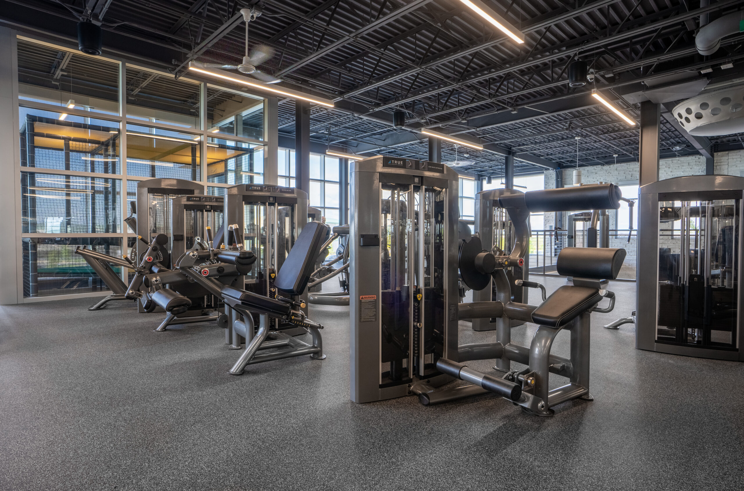 A spacious, modern gym with various strength training machines, large windows for natural light, and a clean, open floor plan. No people are present, and the area looks tidy and well-maintained.