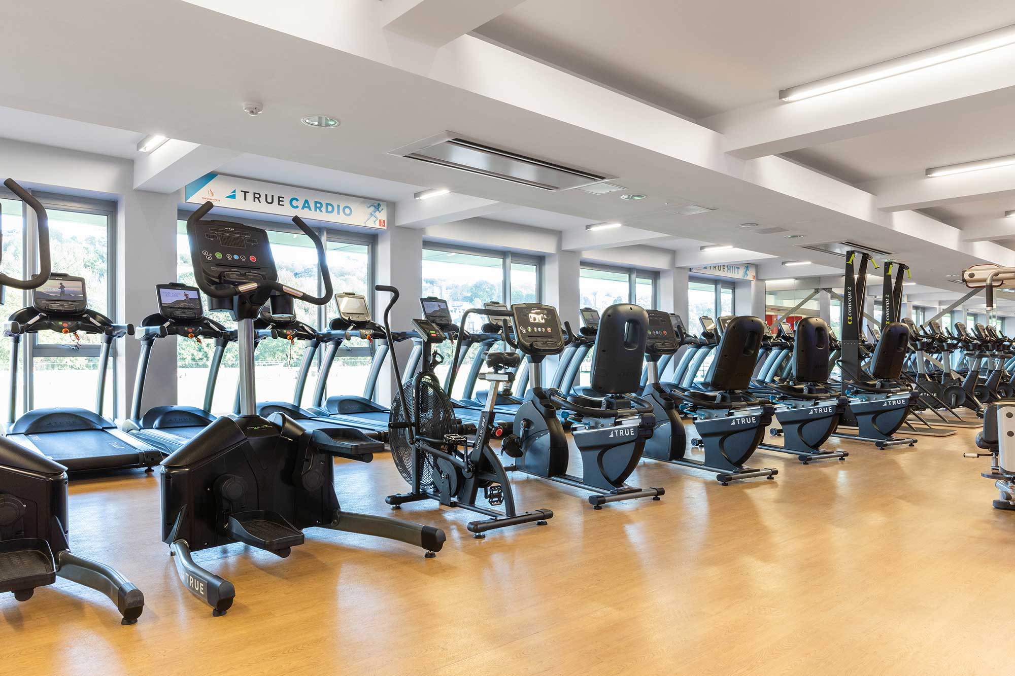 A bright, spacious gym with large windows, featuring rows of treadmills, exercise bikes, and elliptical machines on a light wooden floor under overhead lights.