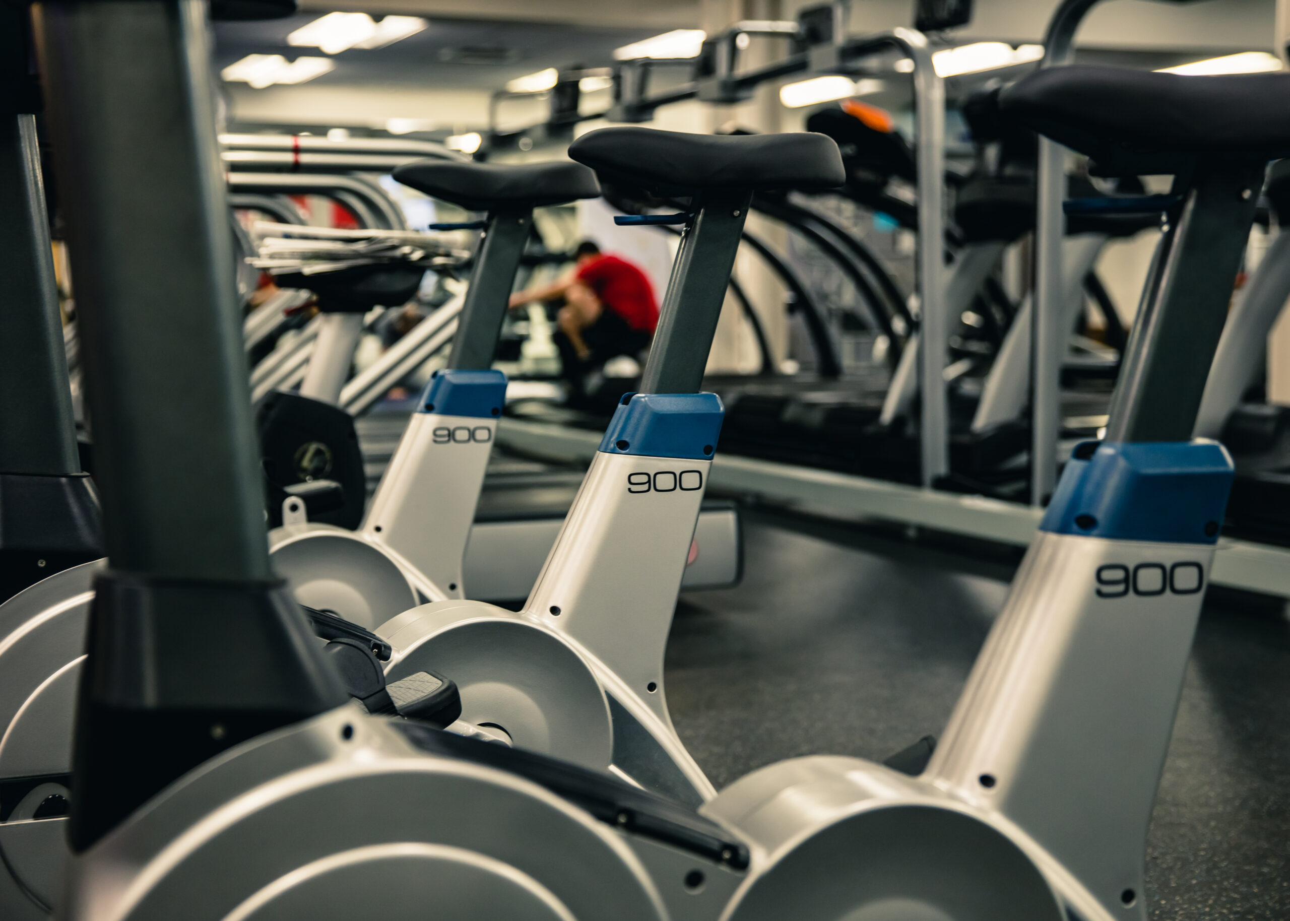 A row of stationary exercise bikes in a gym, with treadmills and other fitness equipment in the background. A person in red is seen kneeling or stretching near the machines.