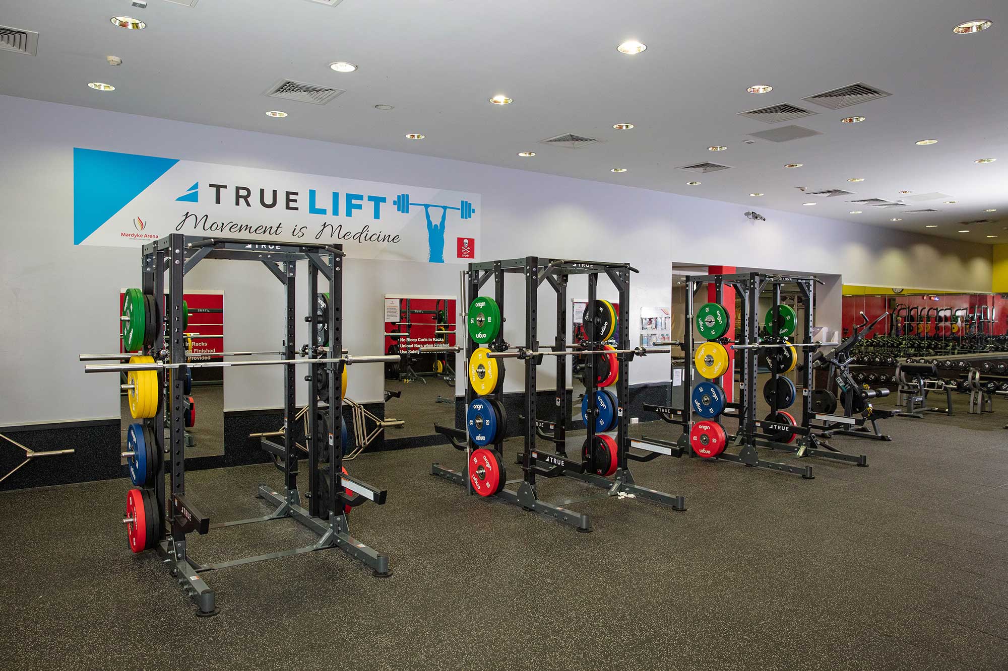 A spacious gym with multiple squat racks holding colorful weight plates, benches, and various gym equipment. A wall sign reads “TRUELIFT Movement is Medicine.” The gym is well-lit and organized.