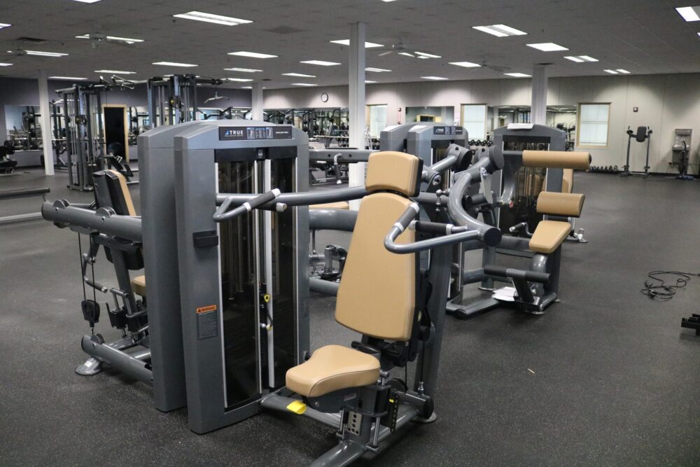 A spacious gym interior with multiple strength training machines, including seated chest press and shoulder press machines. The room has mirrored walls, rubber flooring, and overhead fluorescent lighting.