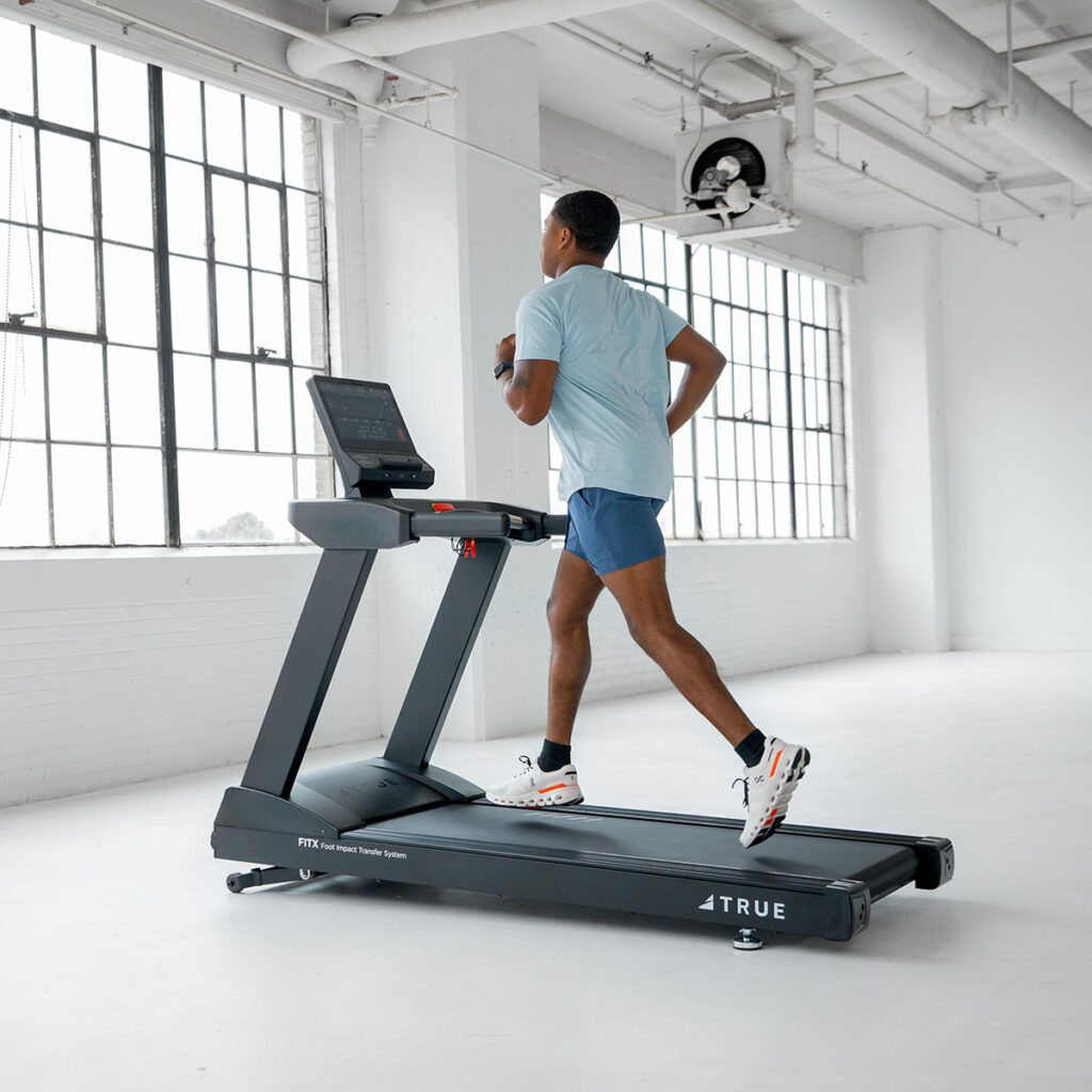 A person in a light blue shirt and shorts is running on a treadmill in a bright, spacious room with large windows and white walls.