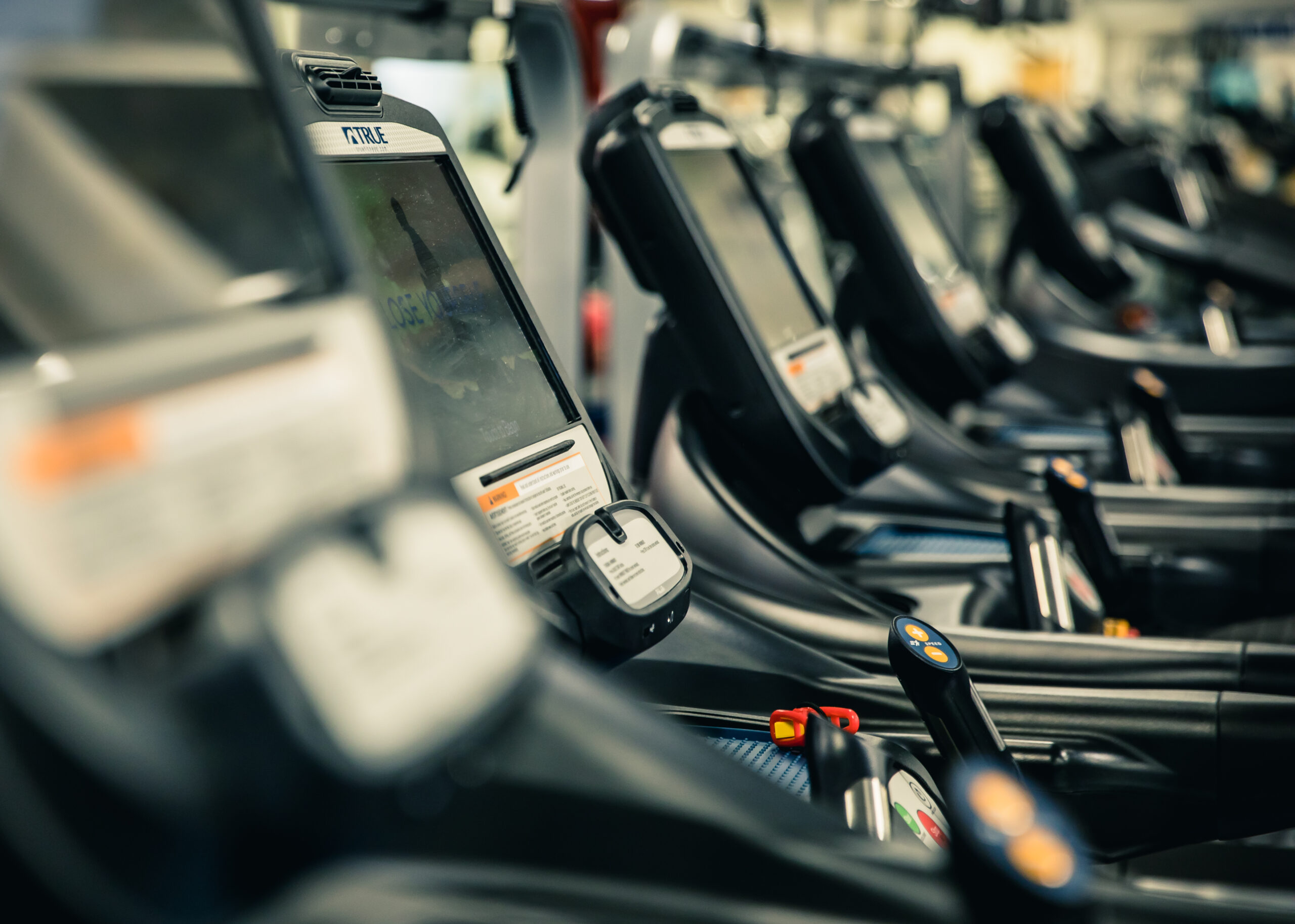 A row of treadmills with digital screens and control panels in a gym, photographed from a side angle with a shallow depth of field.