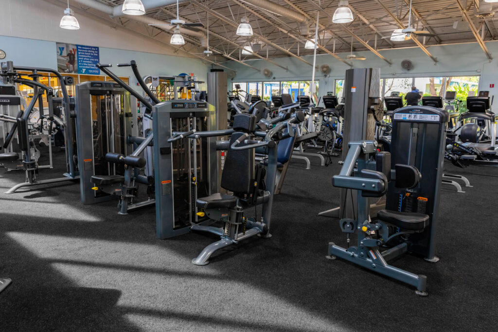 A gym with various exercise machines and equipment arranged on a black carpet, with natural light coming through large windows and several overhead lights brightening the space.
