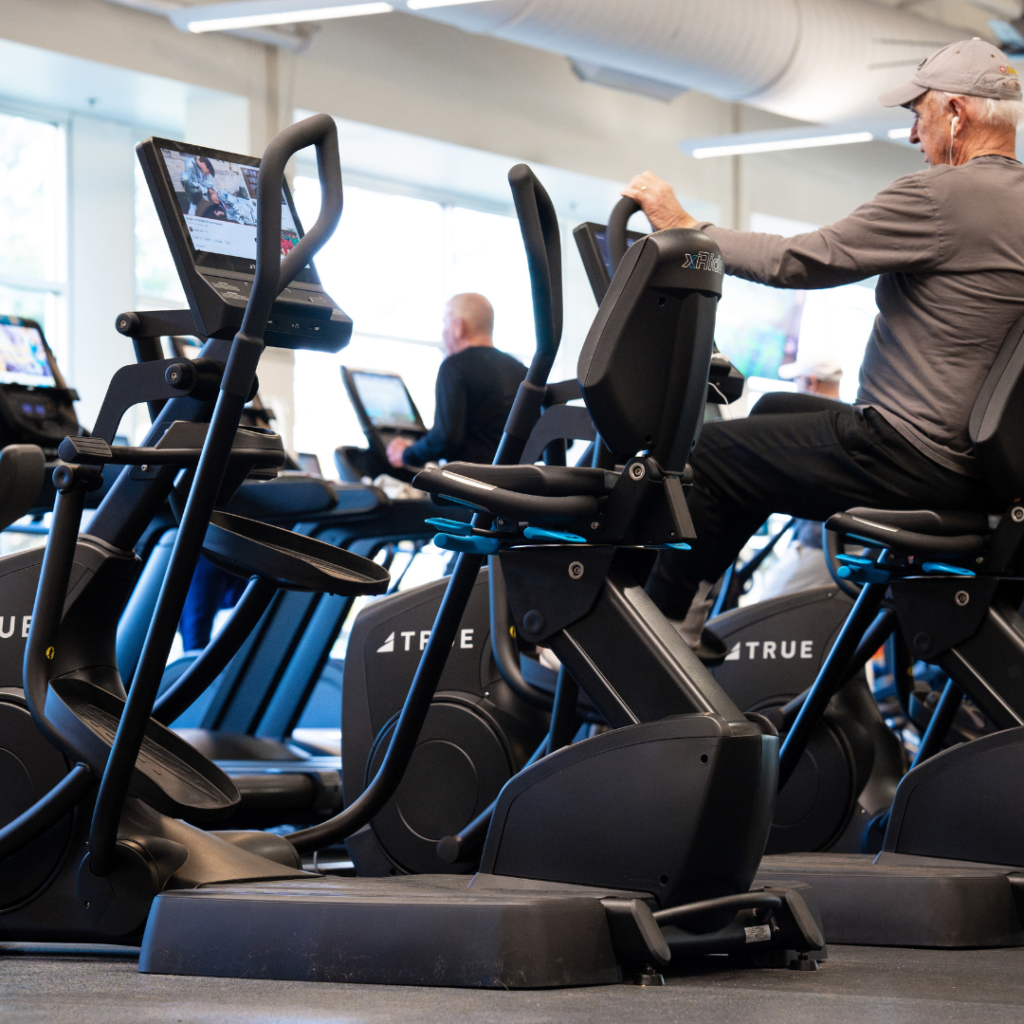 Older man wearing a cap uses a stationary recumbent bike at a gym. Other people are also exercising on similar machines in the background, with TV screens attached to each machine.