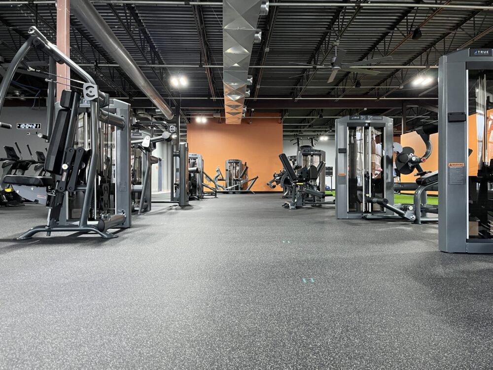 A spacious gym with various weight machines and fitness equipment, featuring rubber flooring and an orange accent wall. The area is well-lit with overhead lights and has an industrial-style exposed ceiling.
