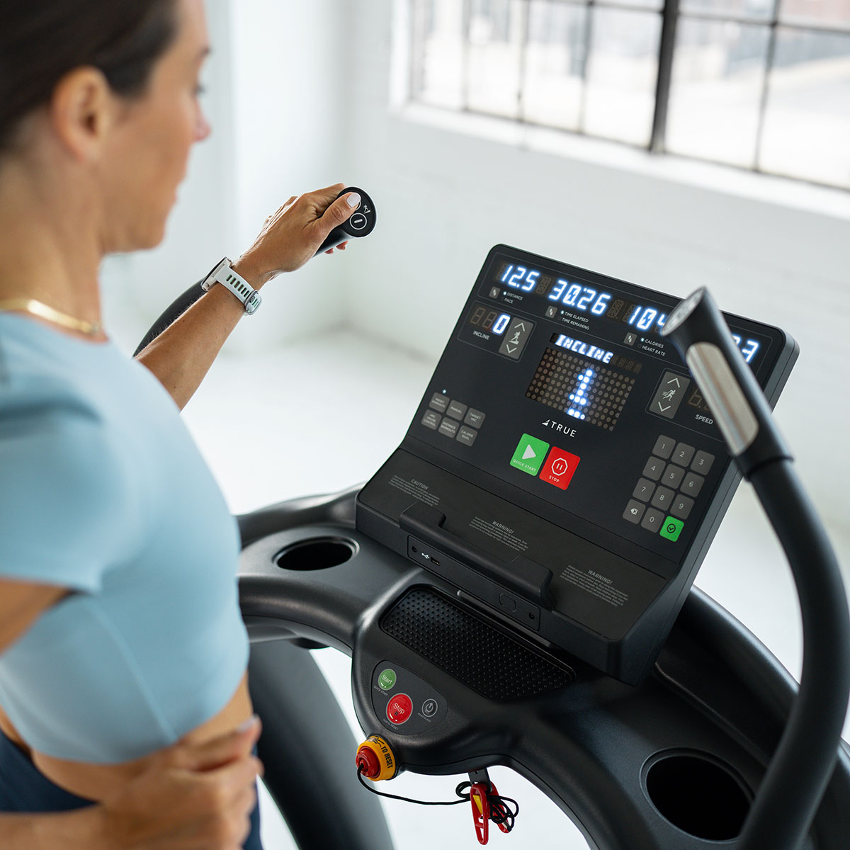 A woman in athletic wear uses a treadmill, holding the handle and adjusting settings on the console, which displays time, speed, distance, incline, and other workout information. Bright daylight streams through a window.