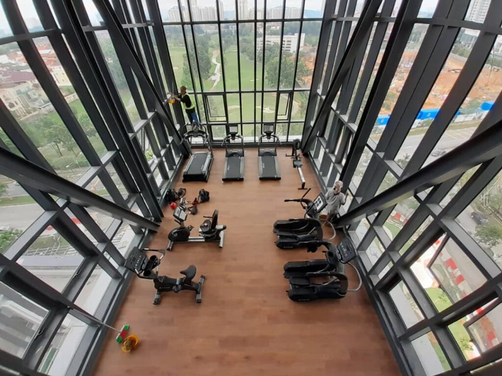 A modern gym in a high-rise building with floor-to-ceiling windows, featuring treadmills, exercise bikes, and elliptical machines on a wooden floor, with views of a city and green park outside.