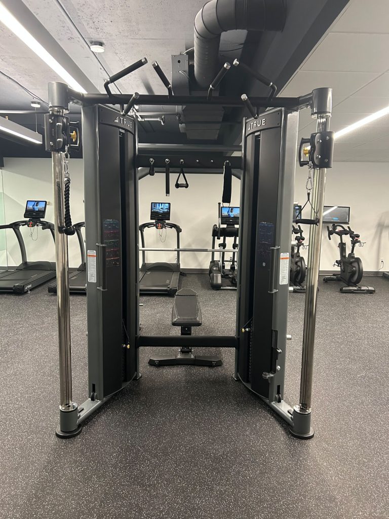 A modern gym with a dual cable machine in the foreground, a workout bench in the center, and treadmills and exercise bikes with screens in the background on a black rubber floor.