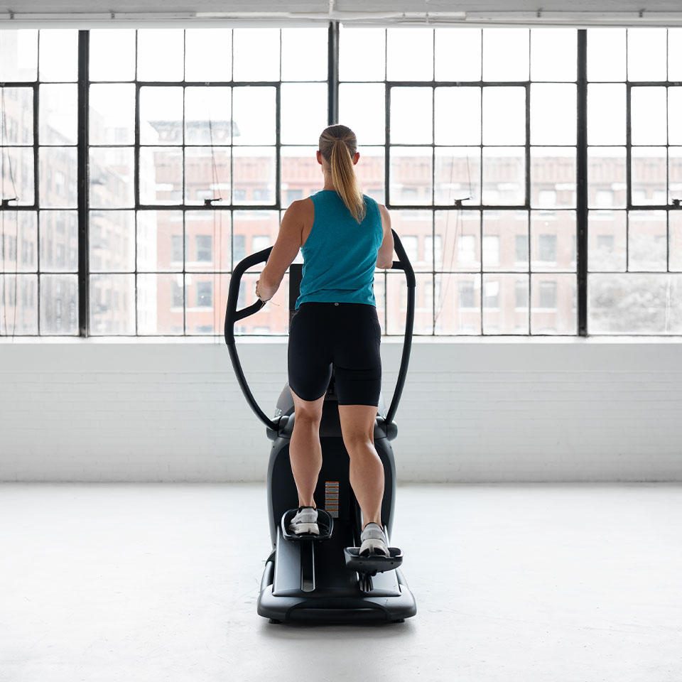 A person with a ponytail wearing a blue sleeveless top and black shorts is exercising on an elliptical machine in a bright room with large windows overlooking city buildings.