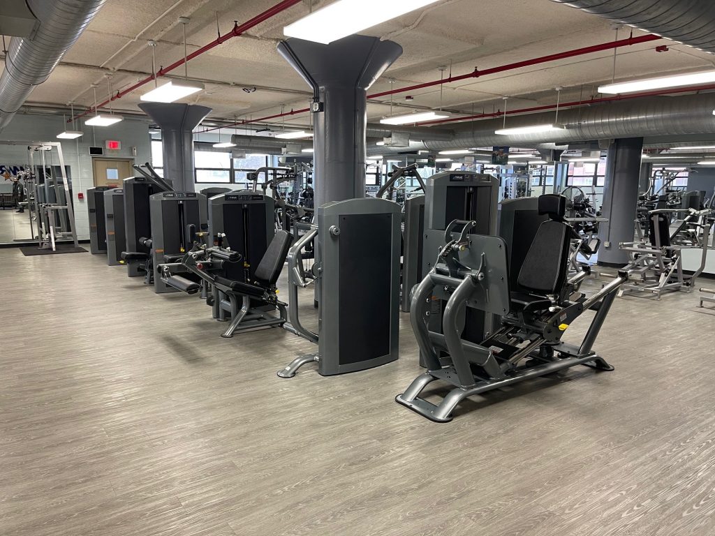 A spacious, modern gym with several black and gray weight machines arranged in rows on a light wood floor, large pillars, mirrors, and windows providing natural light.