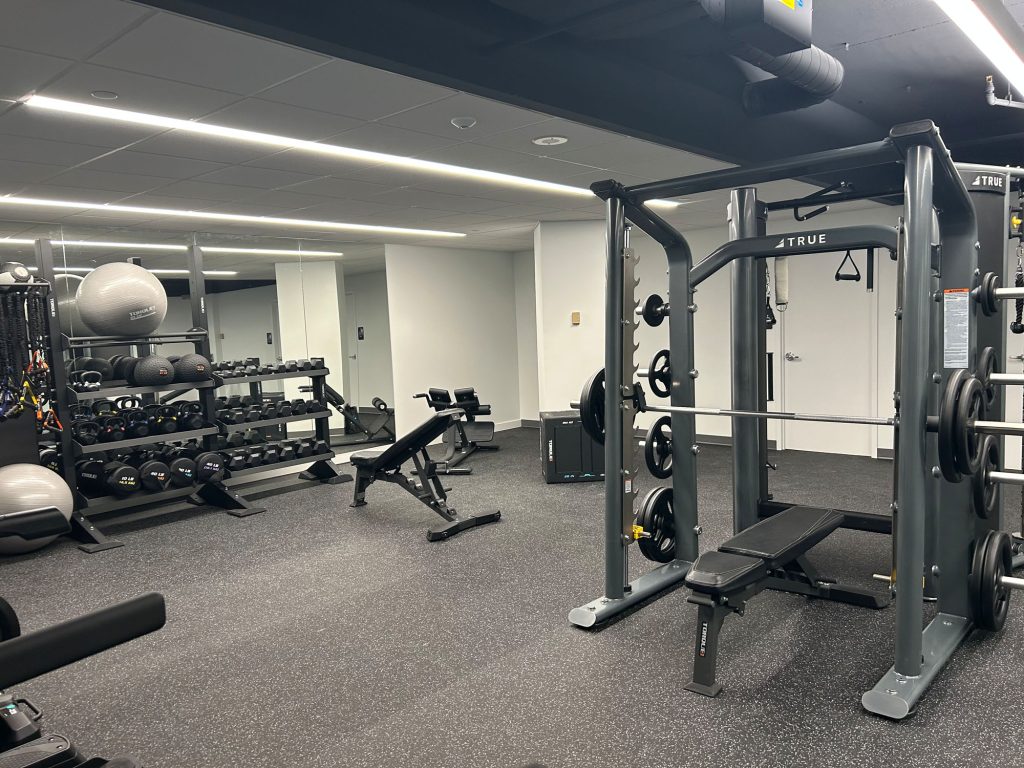 A modern gym with weight racks, benches, a Smith machine, exercise balls, and mirrors along the wall. The space is well-lit, organized, and furnished with various strength training equipment.