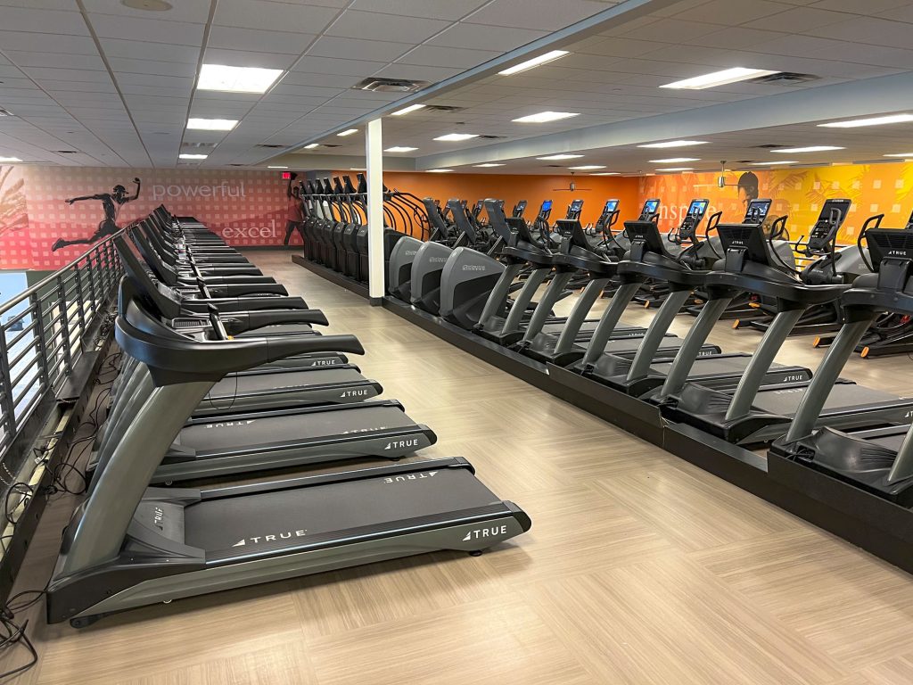 Rows of treadmills and elliptical machines in a brightly lit gym, with motivational words like “powerful” and “excel” on the walls in the background. The floor is covered with light brown tiles.