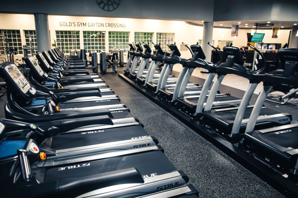 A row of treadmills in a brightly lit gym, with the sign Golds Gym Gayton Crossing visible on the far wall, and other exercise equipment arranged in the background.