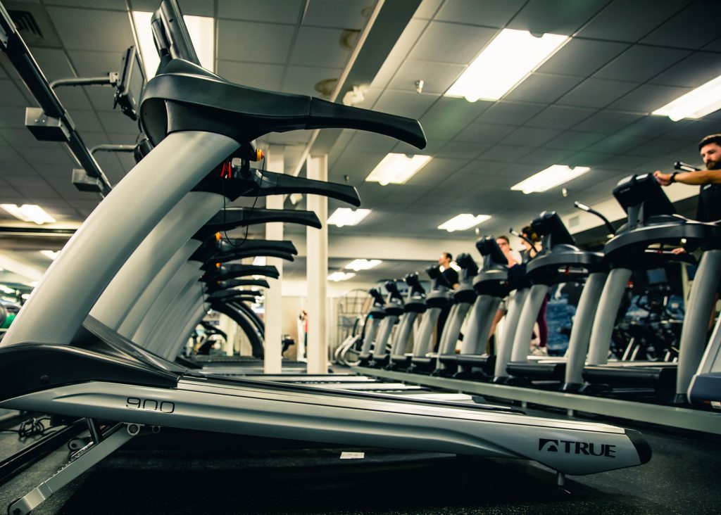 A row of treadmills in a brightly lit gym, with a few people exercising on them in the background. The image is taken from a low angle, focusing on the side of the treadmills.