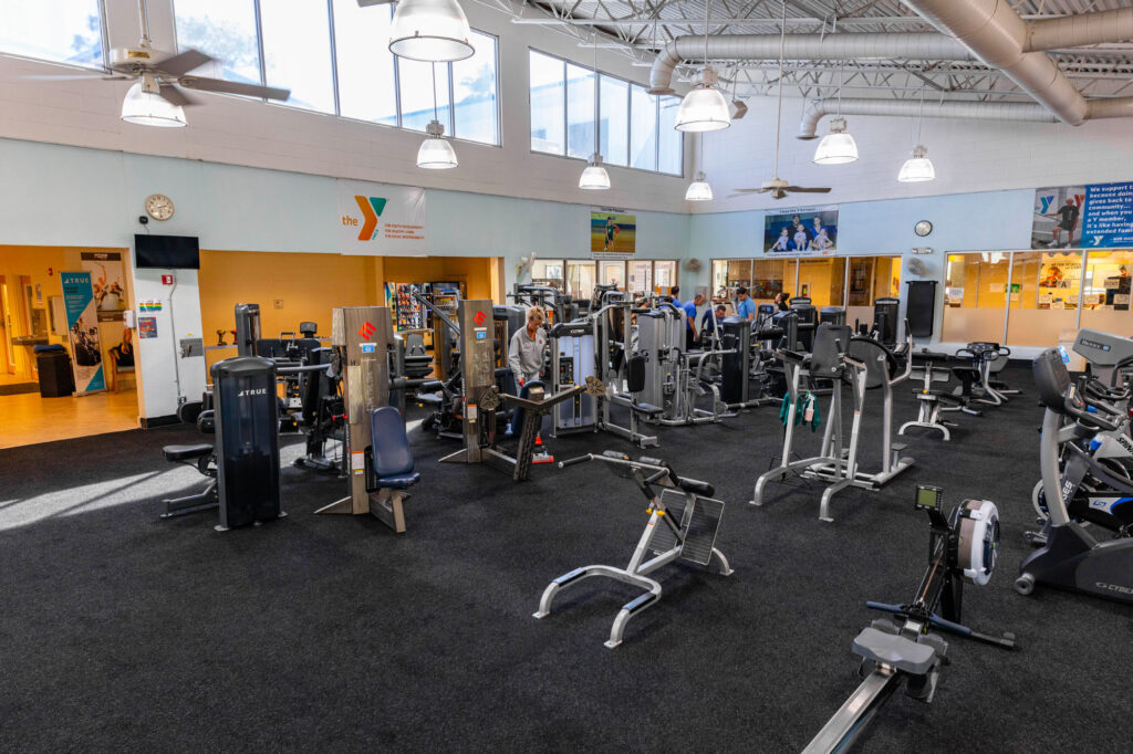 A spacious gym with various exercise machines, including treadmills, weight machines, and bikes, on a black carpeted floor. Large windows, bright overhead lights, and a few people working out are visible.