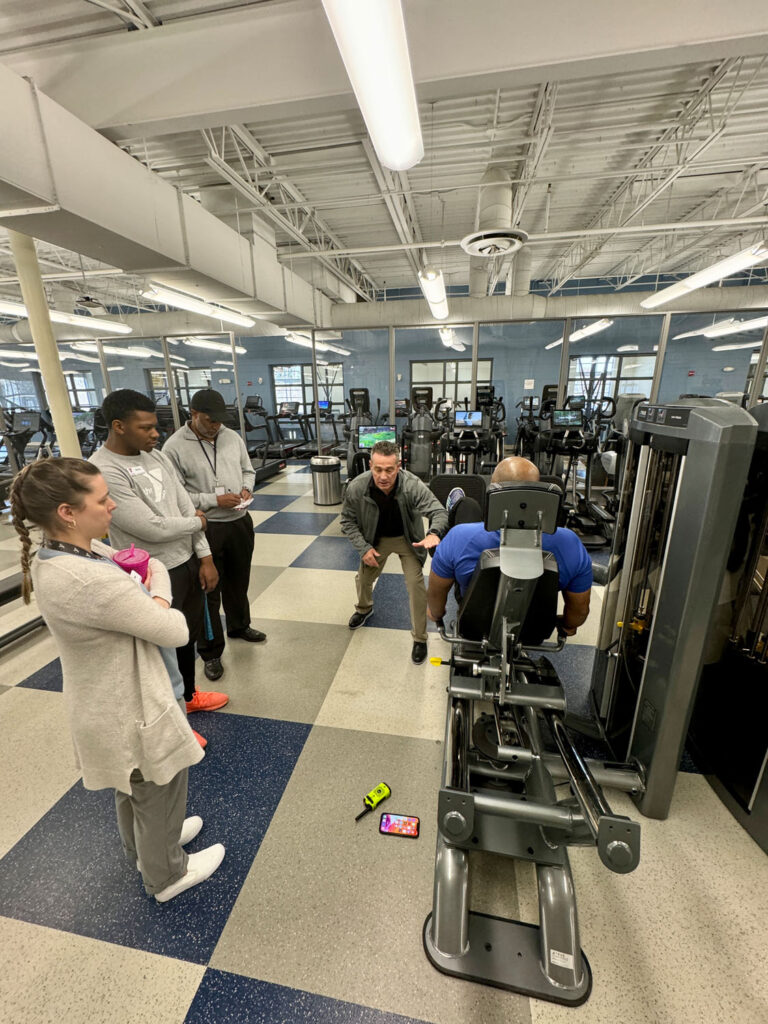A group of five people stands around a seated man using a leg press machine in a spacious gym. One person demonstrates proper technique while others observe attentively. Fitness equipment is visible in the background.