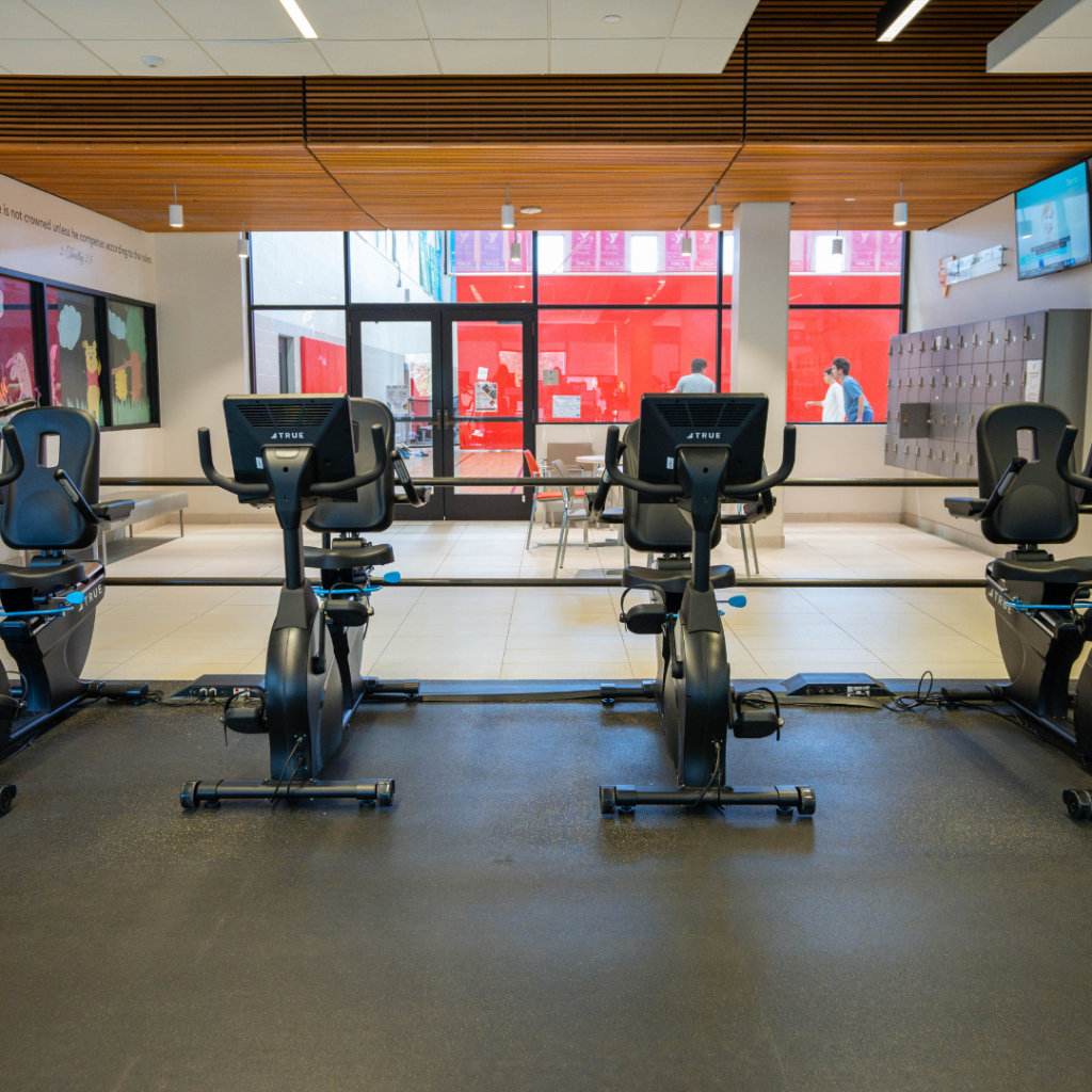 Three stationary exercise bikes face large windows in a modern, bright gym. The space features lockers on the right wall, televisions, and a few people visible through the windows in a red-accented hallway.