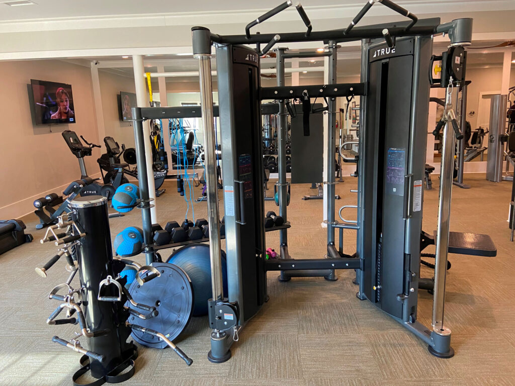 A gym with various fitness equipment, including a multi-functional cable machine, dumbbells, kettlebells, weight plates, resistance balls, and exercise bikes; a TV is mounted on the wall in the background.