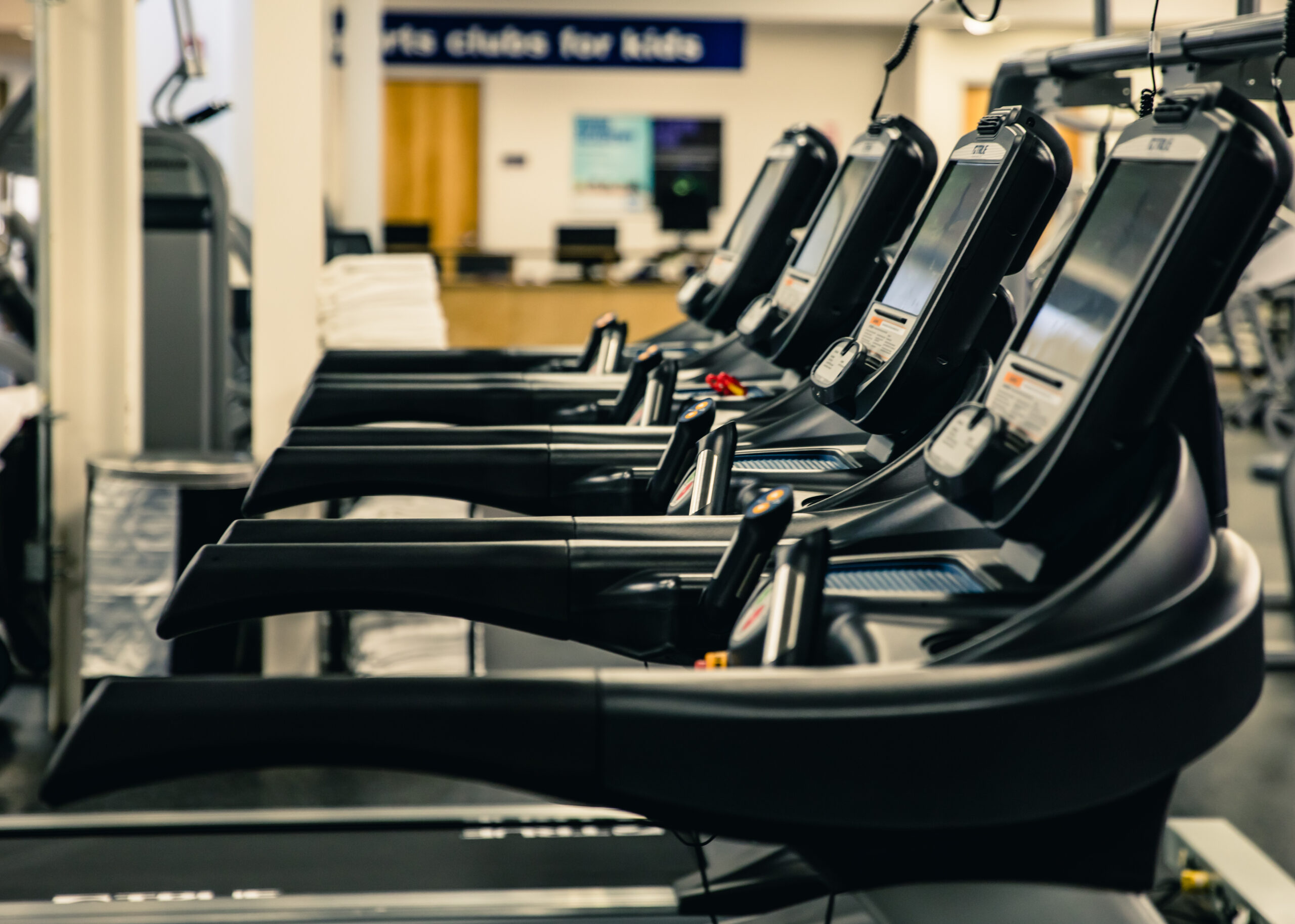 A row of treadmills in a gym, facing the same direction. The gym is well-lit, and there are towels stacked on a counter in the background. The overall scene appears clean and organized.