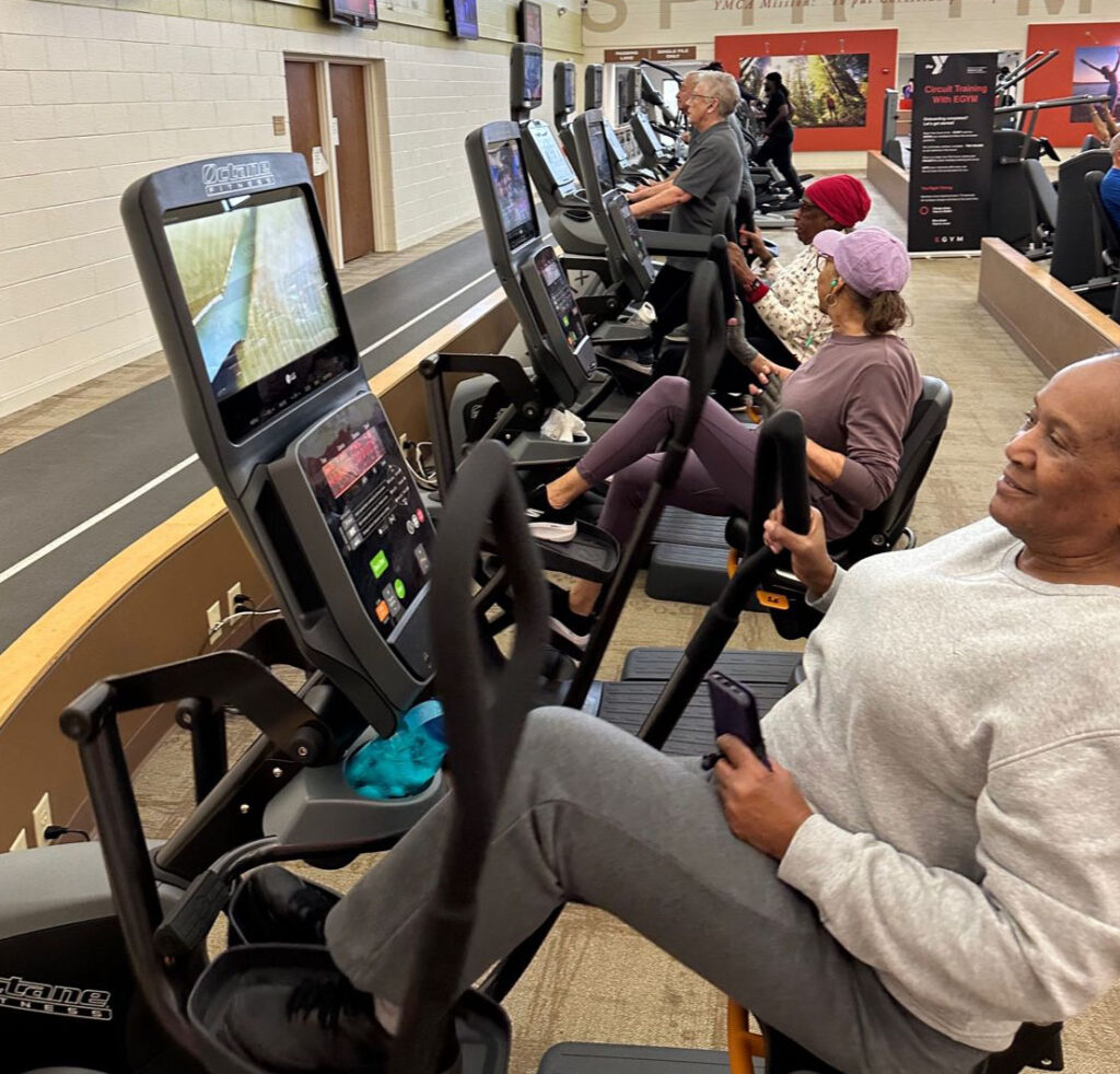 Several adults exercise on recumbent bikes at a gym, using screens on the machines. A walking track and treadmills with more people are visible in the background. The atmosphere appears active and focused.