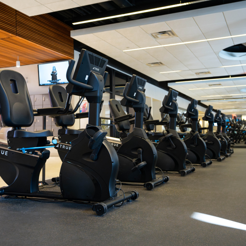 A row of stationary recumbent exercise bikes is lined up in a well-lit, modern gym with large windows and ceiling lights. The gym appears clean and organized, with other equipment visible in the background.