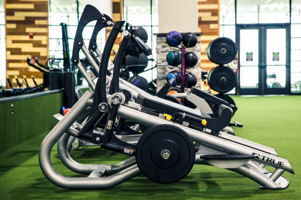 Row of weight machines lined up on green turf in a modern gym, with colorful weight plates and medicine balls stacked in the background. Daylight streams in through large glass doors and windows.