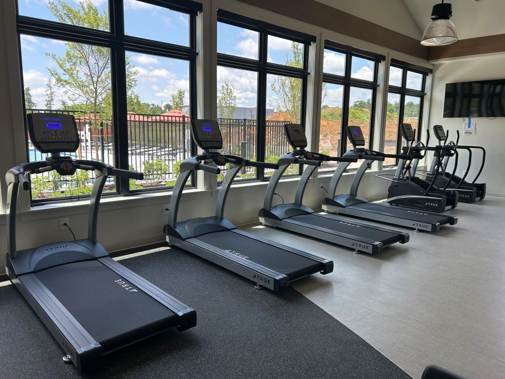 A row of five treadmills is lined up in a bright gym with large windows, allowing natural light to fill the room. Trees and a pool area are visible outside. A TV is mounted on the wall to the right.