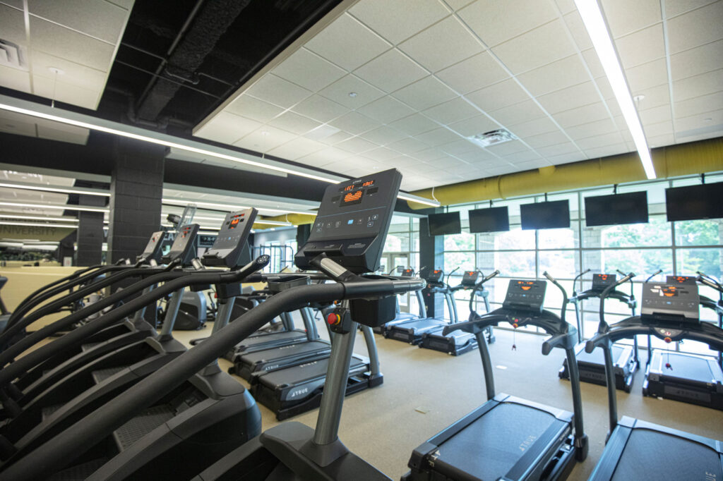 Rows of treadmills are arranged in a spacious, well-lit gym with large windows letting in natural light. Overhead lights and monitors are visible, creating a modern fitness environment.