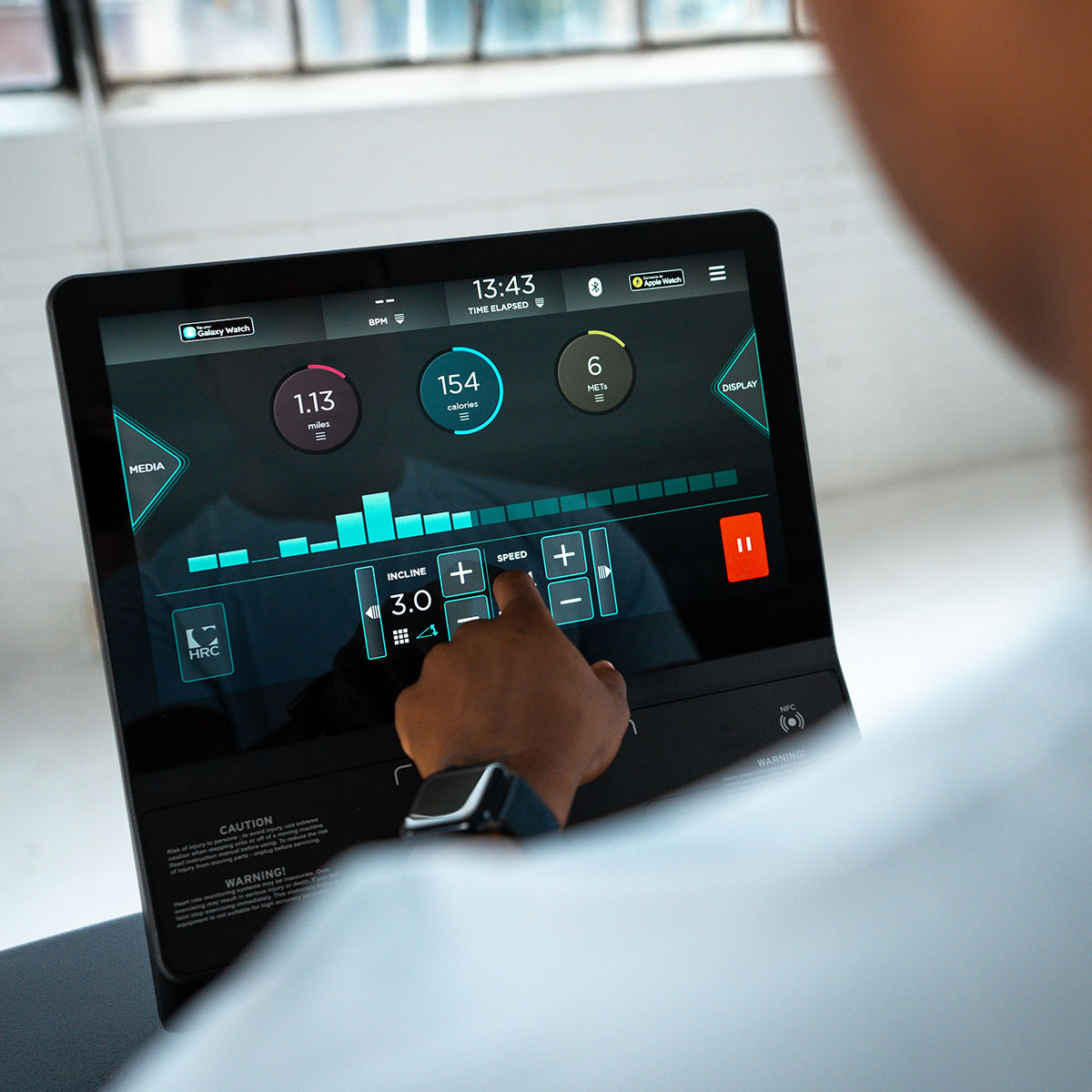 A person uses a touchscreen on exercise equipment, adjusting settings like incline and speed. The display shows workout data such as calories burned, distance, and time elapsed. The person wears a smartwatch.