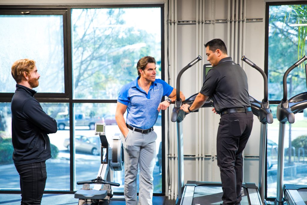 Three men in activewear stand and talk by exercise machines in a gym with large windows. One man stands on a machine, while the other two listen and interact in a bright, modern setting.