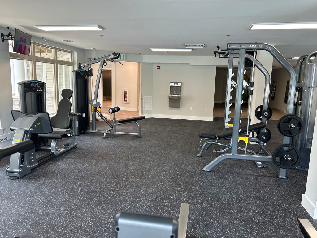 A modern gym with various exercise equipment, including weight machines, benches, and free weights. The room has rubber flooring, large windows, and bright lighting. A TV is mounted in the corner, and a water fountain is visible in the back.