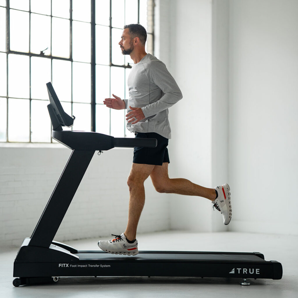 A man wearing a gray long-sleeve shirt and black shorts runs on a treadmill in a bright, sunlit room with large windows.