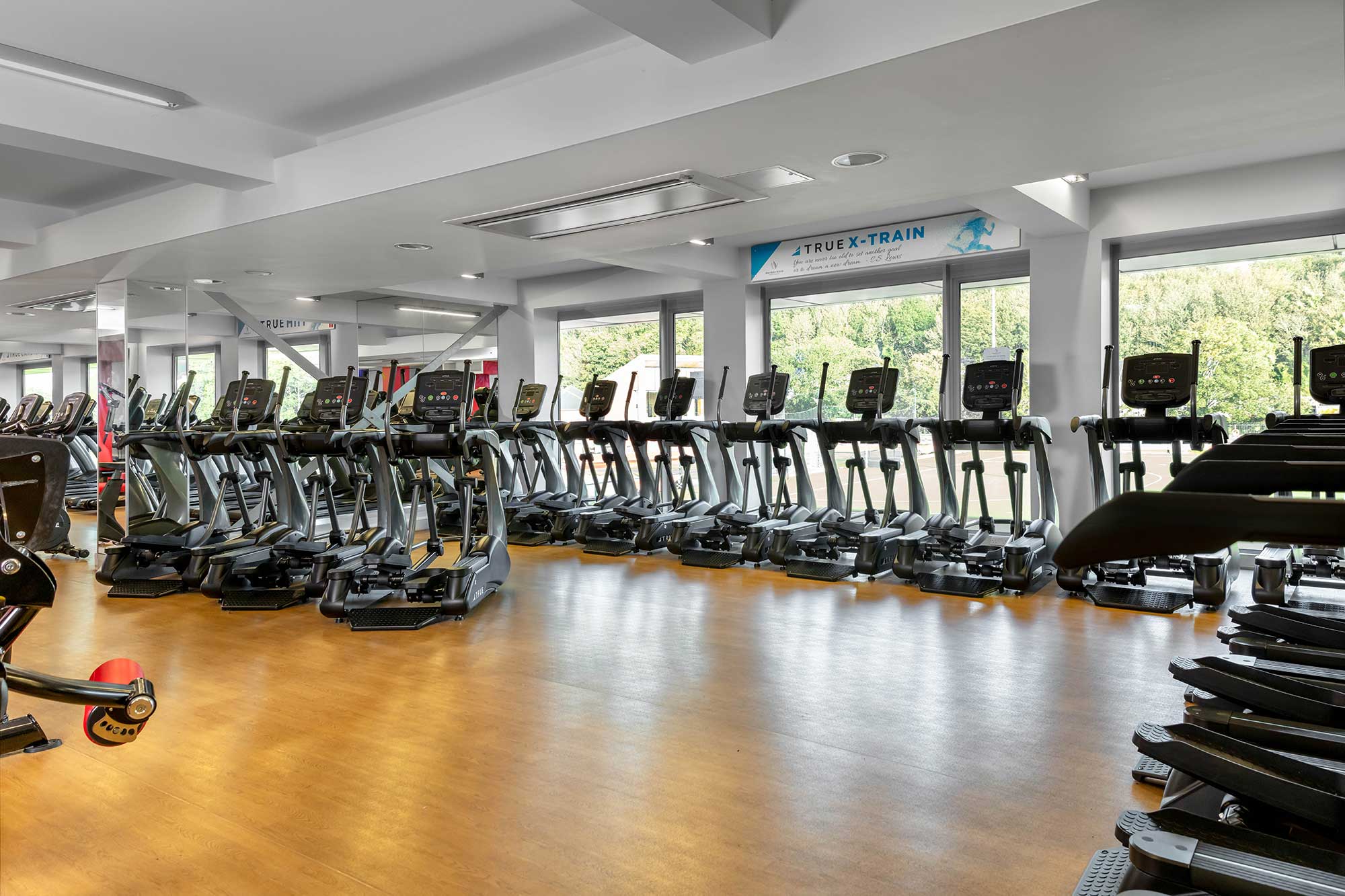 A gym with rows of treadmills and elliptical machines lined up by large windows, letting in natural light. The floor is wooden, and the space appears clean and modern.