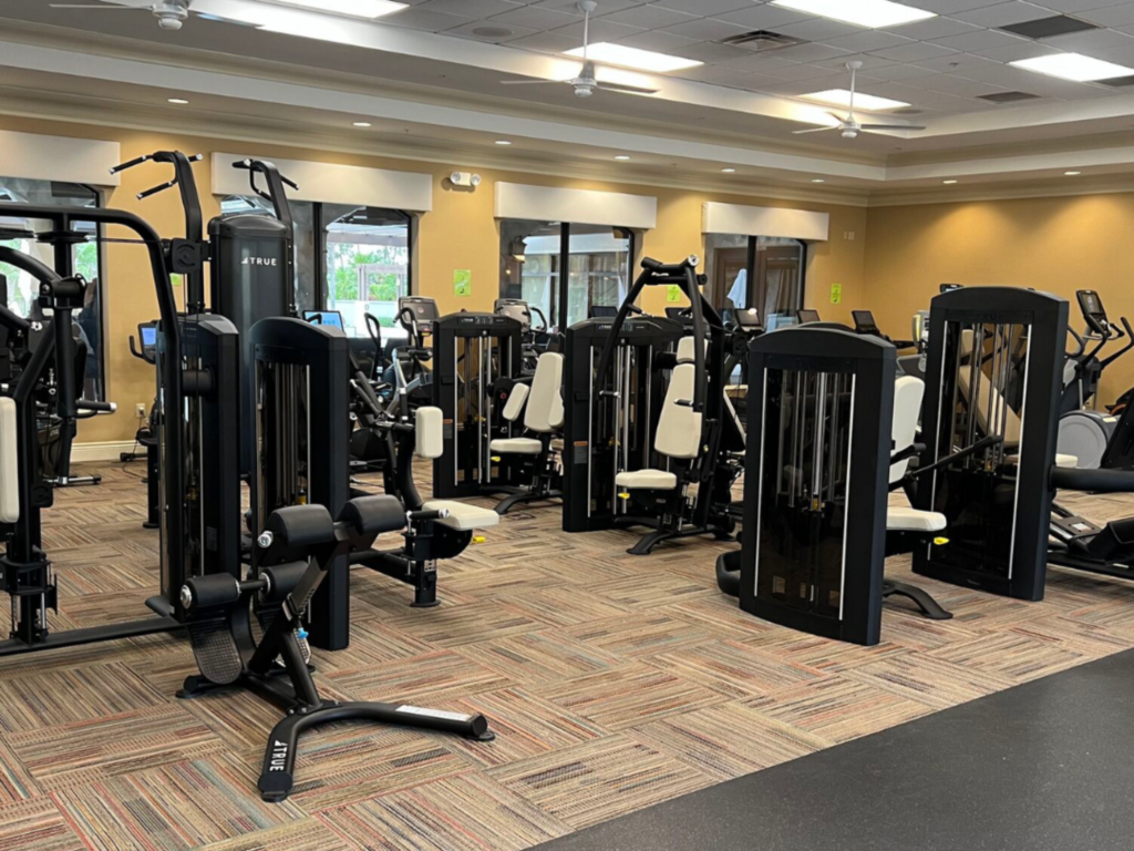 A spacious gym with various strength training machines and cardio equipment, all featuring black frames and white seats, arranged neatly on a carpeted floor under bright ceiling lights.