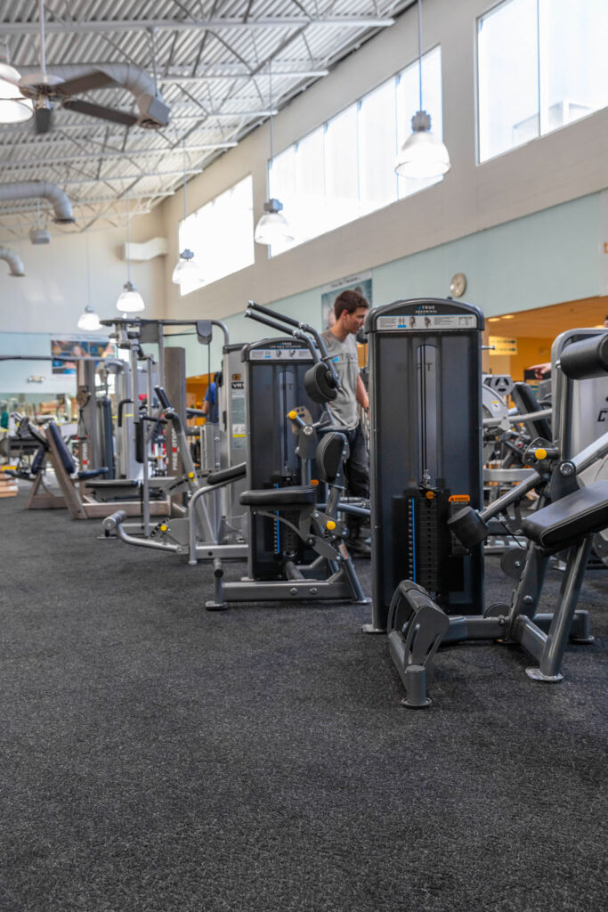A man stands near weight machines in a spacious, well-lit gym with large windows, high ceilings, and various exercise equipment on black carpeted flooring.