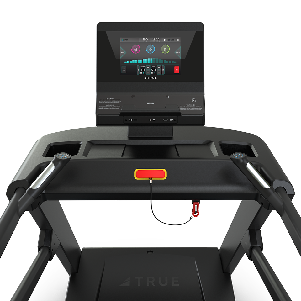 Close-up view of a treadmill control panel with a digital display, showing workout stats, and a red safety key attached by a cord. The TRUE brand logo is visible on the machine.