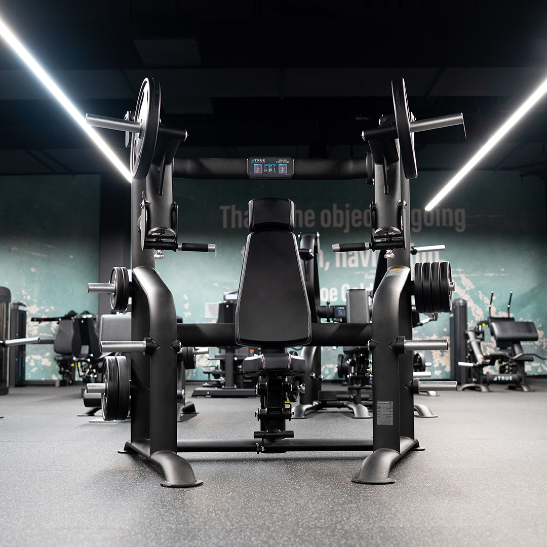 A gym with weight machines, including a seated chest press in the foreground, surrounded by additional fitness equipment under bright overhead lighting.