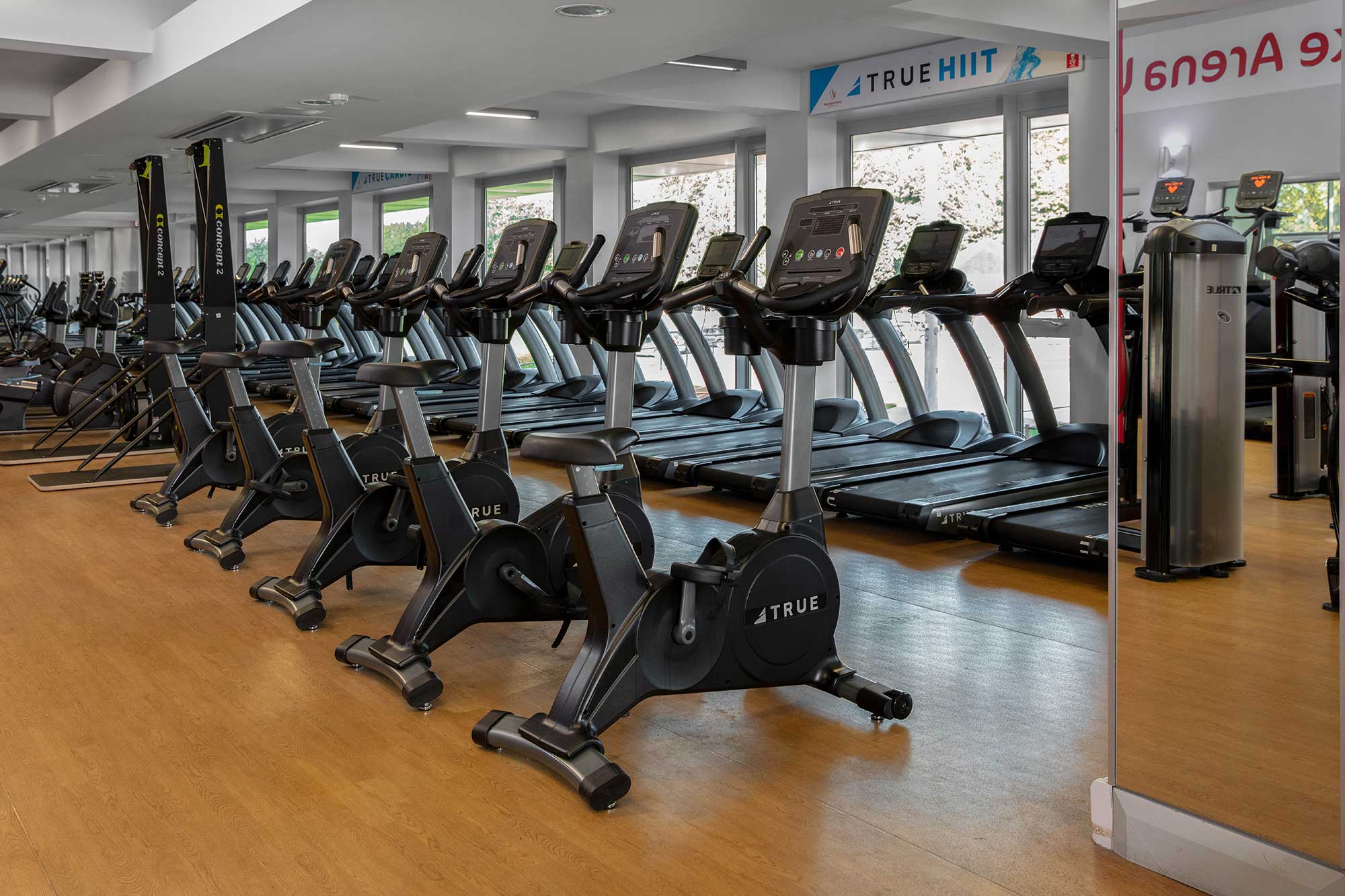 A row of stationary bikes and treadmills are lined up in a modern gym with large windows, mirrors, and wood flooring. The equipment is unused and the space appears clean and well-lit.