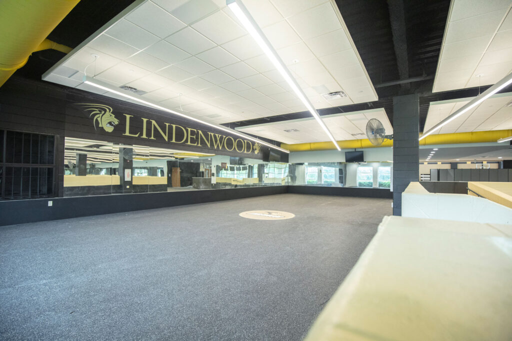 Spacious indoor athletic facility with Lindenwood and a lion logo on the wall, black and yellow accents, large mirrors, and ample overhead lighting.