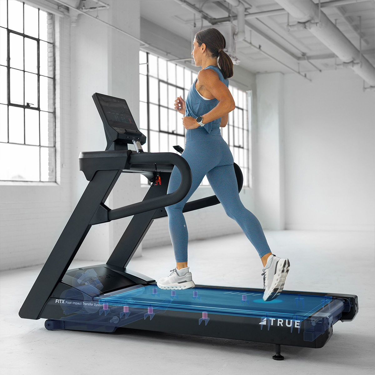 A woman in blue workout clothes runs on a treadmill in a bright, spacious gym with large windows. The treadmill has digital controls and features highlighted inner components in blue.