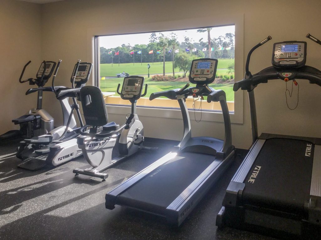 A gym room with exercise equipment, including treadmills, stationary bikes, and an elliptical machine, positioned near a large window that overlooks a green outdoor area with trees and flagpoles.