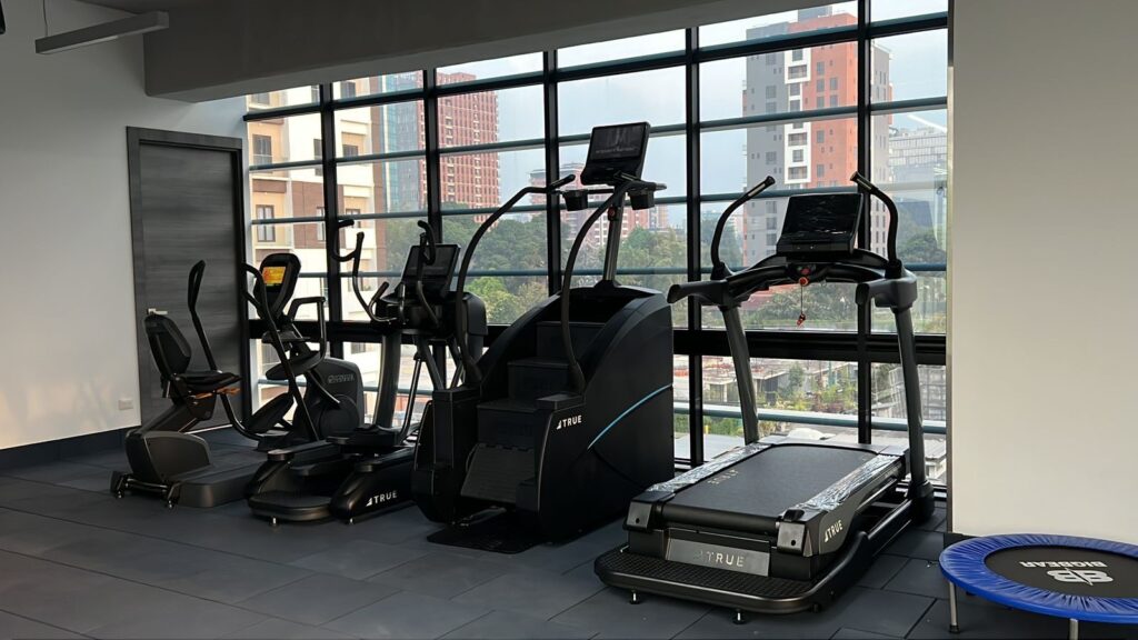 Three modern cardio machines and a small trampoline in a gym with large windows overlooking a cityscape with tall buildings and trees. The room has dark floor tiles and bright natural light.