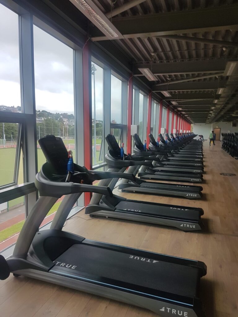 A row of treadmills lined up in a modern gym with large windows overlooking an outdoor sports field. The treadmills are unoccupied and the gym is well-lit with natural light.