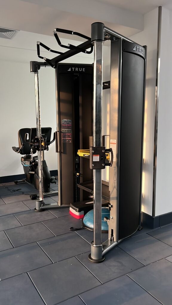 A modern gym machine with pulleys and cables stands on a tiled floor near a recumbent bike. The equipment is black with chrome details and labeled TRUE. Sunlight shines on the scene.