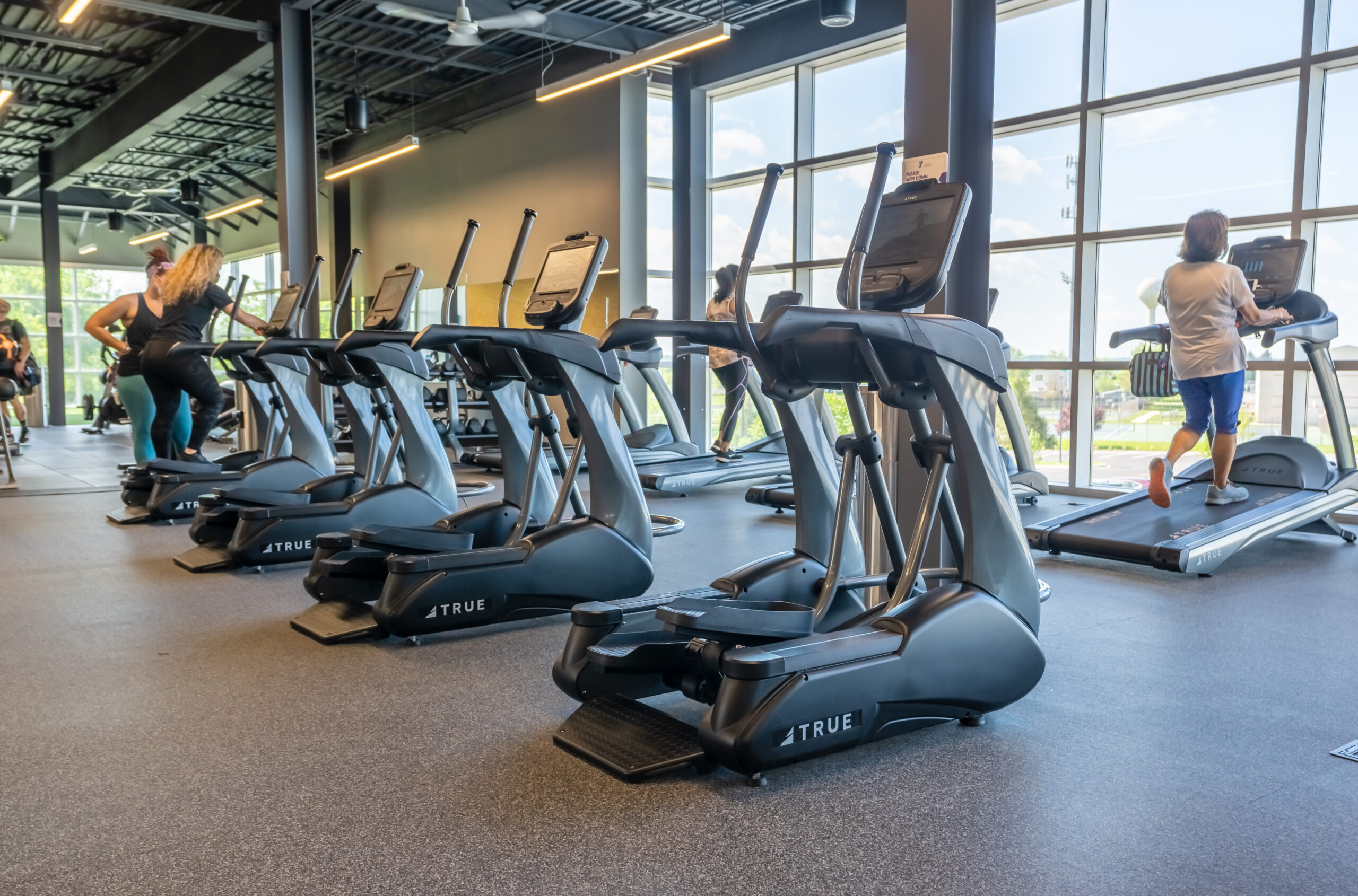 A row of empty elliptical machines in a modern gym with large windows, while people exercise on treadmills in the background. Bright daylight streams in through the windows.